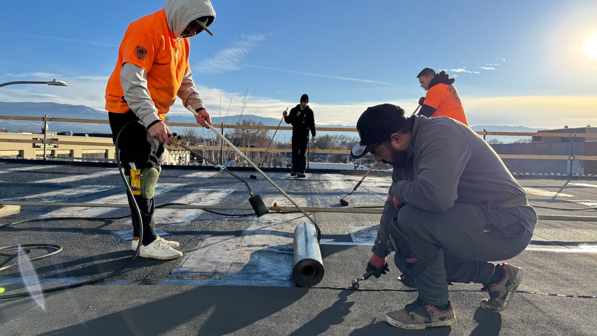 Two roofers torching a new roll of membrane down over the old flat roof, a third crew member behind them