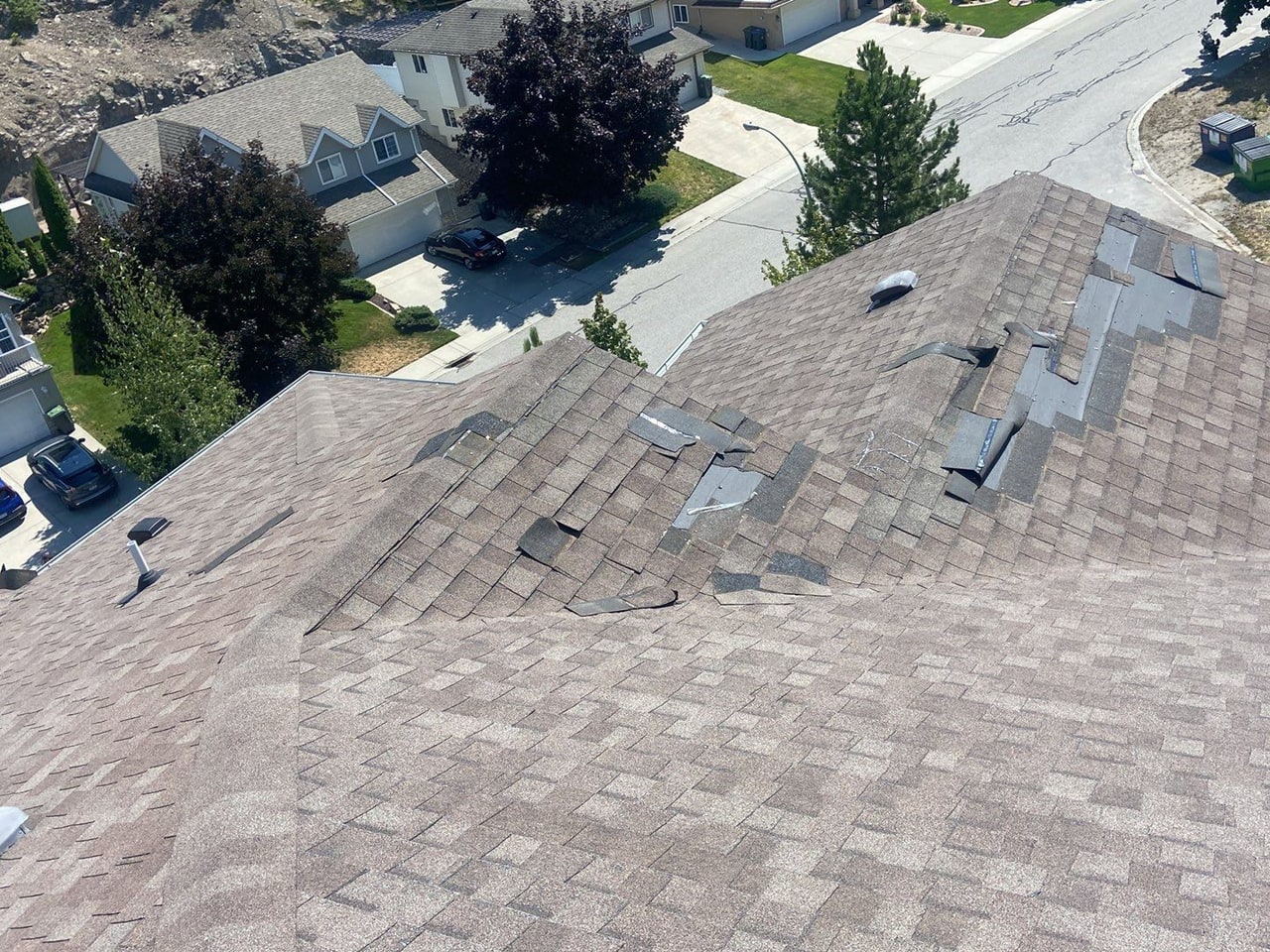 A shingle roof from above after a windstorm: shingles lifted and torn off across the slope, the street below