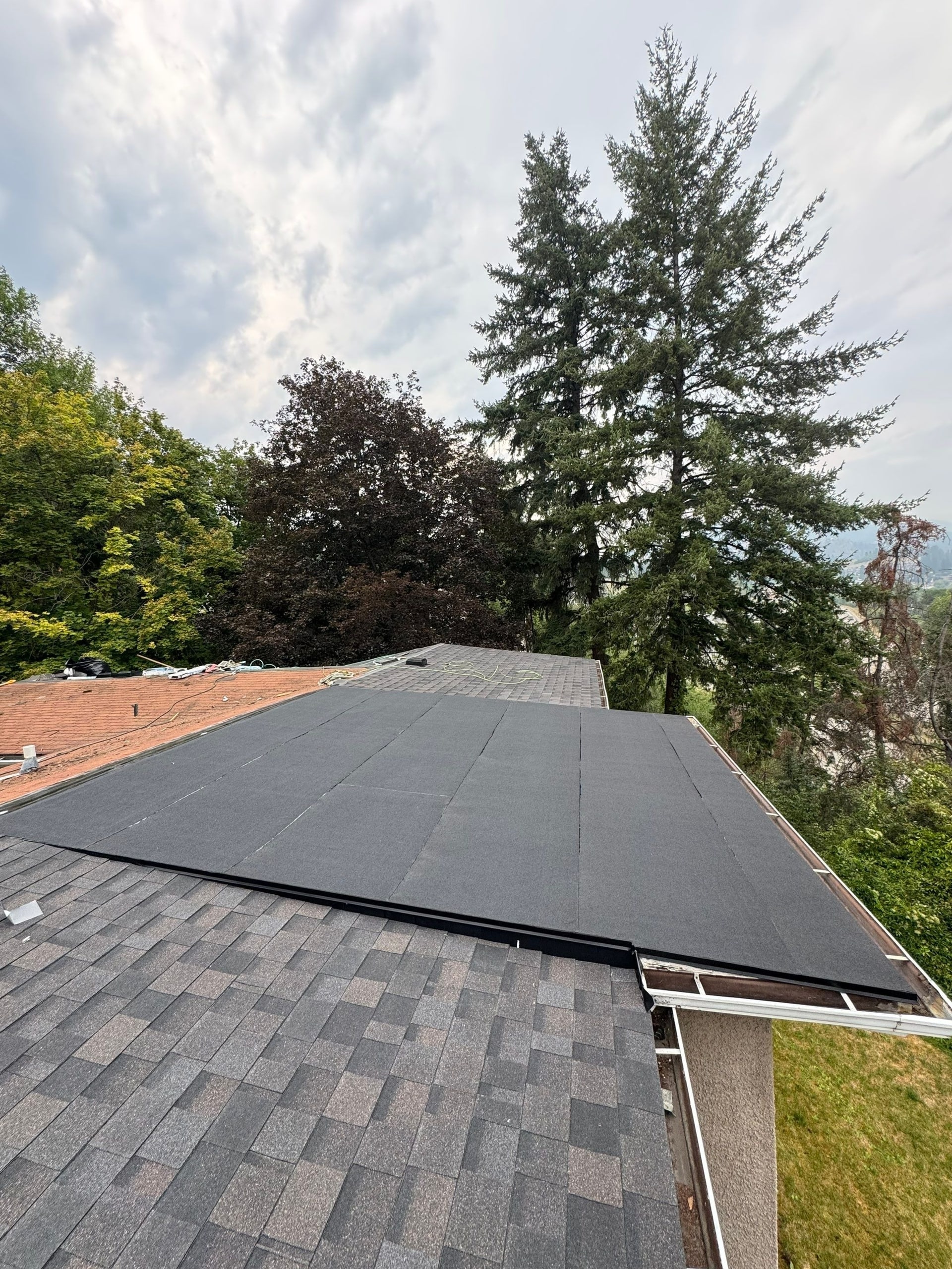 A low-slope part of a roof in new black membrane, new charcoal shingles around it, trees and the valley beyond